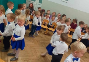 Dzieci z najmłodszej grupy spiewają i tańczą w parach, pozostałe starsze dzieci siedzą na ławeczkach i oglądają występ "maluszków"
