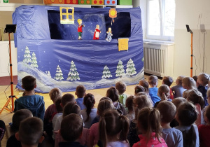 Dzieci siedzą na ławeczkach i oglądają przedstawienie z udziałem lalek i aktorów