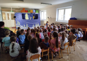 Dzieci siedzą na ławeczkach i oglądają przedstawienie teatralne