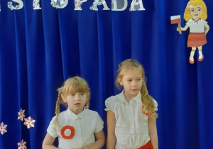 Dziewczynki ubrane na biało czerwono mówią wiersze na tle dekoracji święta 11 listopada