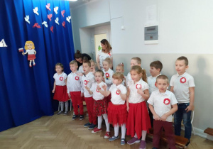 Dzieci ubrane na biało czerwono mówią wiersze na tle dekoracji święta 11 listopada