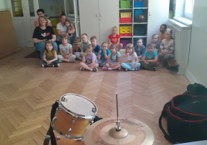 Dzieci z grupy IV słuchają o instrumentach perkusyjnych siedząc na podłodze.