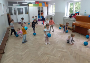 Dzieci z piłkami w rękach stoją na sali i wykonują ćwiczenia oraz uczestniczą w zabawach