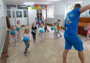 Dzieci z piłkami w rękach stoją na sali i wykonują ćwiczenia oraz uczestniczą w zabawach