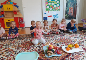 Dzieci siedzą na dywanie i po kolei oglądają i przeliczają warzywa i owoce