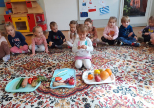 Dzieci siedzą na dywanie i po kolei oglądają i przeliczają warzywa i owoce