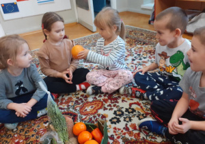 Dzieci siedzą na dywanie i po kolei oglądają i przeliczają warzywa i owoce