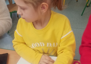 dziewczynka siedzi przy stoliku i wykonuje pracę plastyczną, rysując , wyklejając kolorowym papierem, włóczką, brokatem postać swojej babci lub dziadka