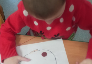 chłopiec siedzi przy stoliku i wykonuje pracę plastyczną, rysując , wyklejając kolorowym papierem, włóczką, brokatem postać swojej babci lub dziadka