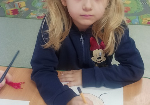 dziewczynka siedzi przy stoliku i wykonuje pracę plastyczną, rysując , wyklejając kolorowym papierem, włóczką, brokatem postać swojej babci lub dziadka