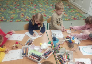 Dzieci siedzą przy stoliku i wykonują pracę plastyczną, rysując , wyklejając kolorowym papierem, włóczką, brokatem postać swojej babci lub dziadka