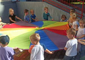 Dzieci trzymają kolorową chustę animacyjną. Śpiewają i wykonują ruchy do piosenek wg pokazu nauczycielki