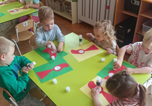 Dzieci siedzą przy stoliku i przyklejają klejem do kartki elementy z kolorowego papieru tworząc postać czerwonego kapturka.