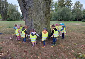 Dzieci stoją wokół drzewa, trzymają się za ręce.