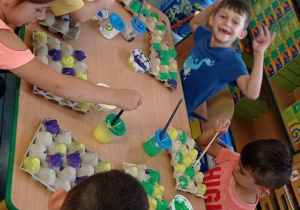 Dzieci siedzą przy stole i malują farbami opakowania tworząc kolorowe puzzle