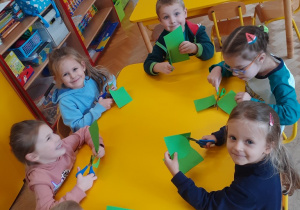 Dzieci z grupy III siedzą przy stoliku i wycinają elementy z zielonej kartki