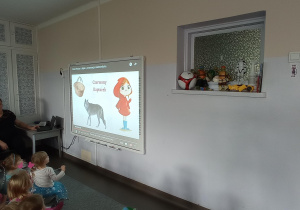 Dzieci siedzą na dywanie i rozwiązują zagadki dotyczące bajek wyświetlane na tablicy interaktywnej