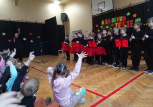 Dzieci ze szkoły tańczą do piosenki, przedszkolaki oglądają występ i naśladują ruchy rękoma w rytm muzyki