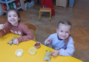 Dzieci siedzą przy stoliku i malują farbami kawałek platonki po jajkach