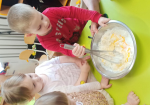 Dzieci stoją przy stoliku na którym jest miska ze składnikami na pączusie, chłopiec miesza składniki