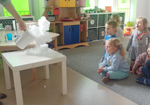 Dzieci siedzą na dywanie i z zaciekawieniem obserwują eksperyment
