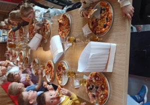Dzieci siedzą przy długim stole i jedzą zrobione przez siebie swoje pizze