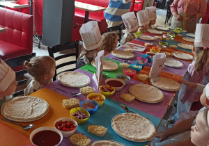 Dzieci siedzą przy stole na głowach mają duże kucharskie czapki. Słuchają opowieści skąd pochodzi pizza. Na talerzach przez dziećmi jest przygotowane ciasto - spód pizzy.