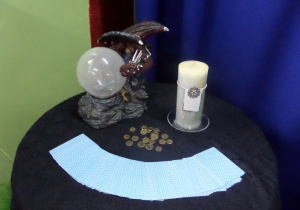 Na stoliku stoją magiczne przedmioty - kryształowa kula, karty, monety i świeca