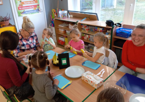 Dzieci siedzą przy stoliku i z pomocą rodziców obierają banany