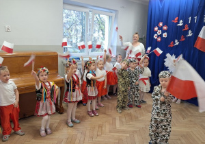 Dzieci śpiewają piosenkę i tzrymają w dłoni flagi wysoko uniesione w górę. Na scenie stoi chłopiec i trzyma dużą flagę Polski