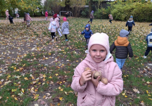 Dziewczynka pozuje do zdjęcia ziemniakami, które znalazła. W tle dzieci zbierające ziemniaki
