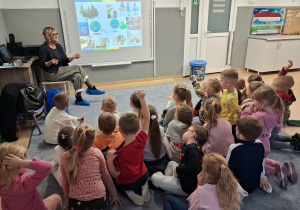 Dzieci siedząc na dywanie, oglądają prezentację multimedialną dotyczącą prawidłowej segregacji śmieci