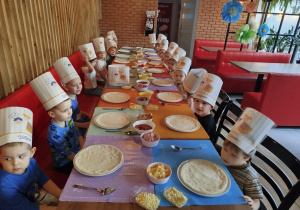 Dzieci siedzą przy wspólnym stole i zaraz zaczną układać składniki na swoich pizzach