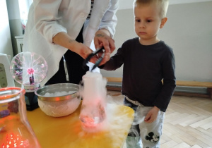 Prowadząca wykonuje eksperymenty z suchym lodem, powiada o nich dzieciom i angażuje dzieci do części zabaw z suchym lodem