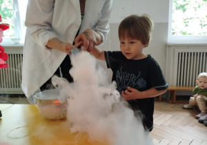 Prowadząca wykonuje eksperymenty z suchym lodem, powiada o nich dzieciom i angażuje dzieci do części zabaw z suchym lodem