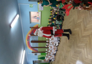 Mikołaj stoi na środku sali i wita się z dziećmi.